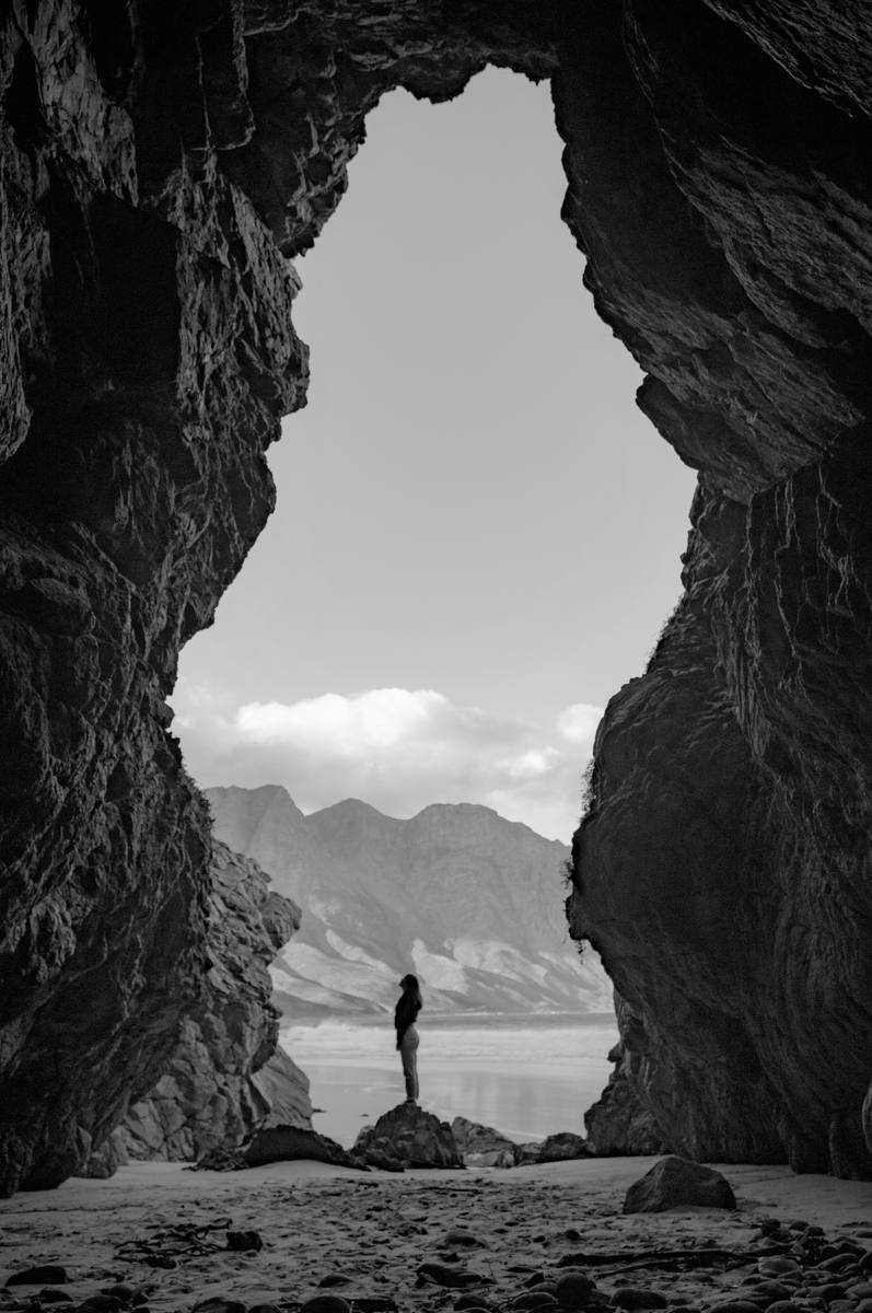 A person standing at the mouth of a sea cave at first light, looking out toward mountains.